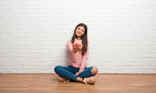 teen sitting against a white wall holding a piggy bank out in front