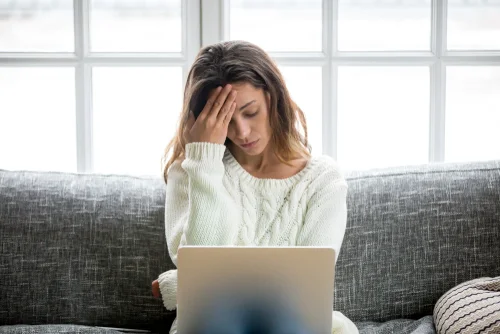 woman sitting on a couch with her hand on her head looking at a computer screen