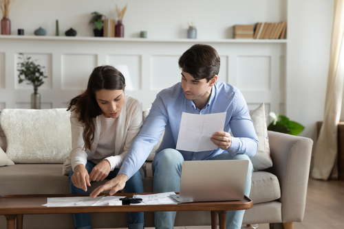 man and woman sitting on couch and looking over paperwork in front of computer screen