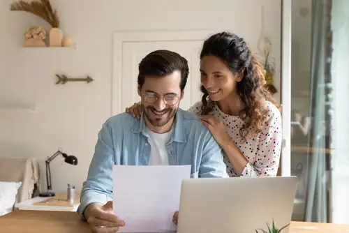 man and woman looking at piece of paper in front of their computer