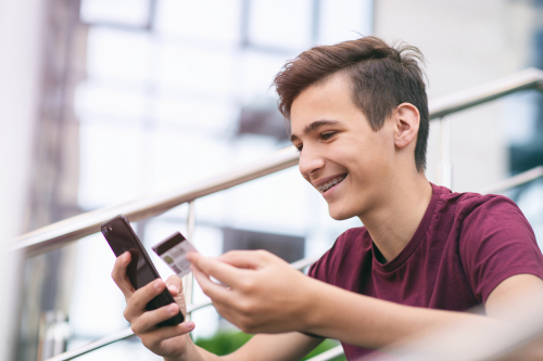 teen sitting outside and looking at his phone while holding a credit or debit card