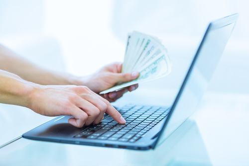 person typing on computer with one hand and holding stack of money in the other