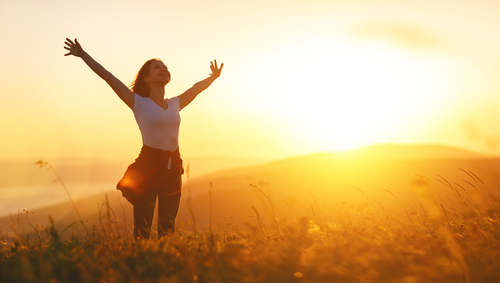 woman standing outside with her arms in the air and sun in the background